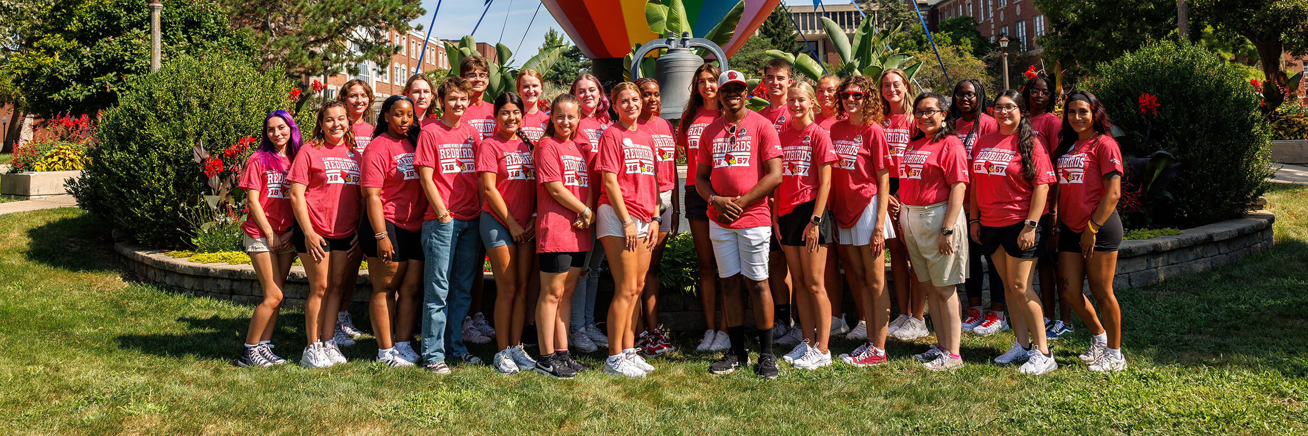 A group of people in matching red T-shirts stand together in front of a large bell and landscaped greenery.